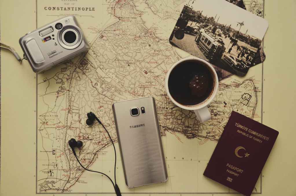 Silver Camera Near Black Coffee in Mug, Silver Samsung Galaxy S7, Turkey Passport, and Black Earbuds