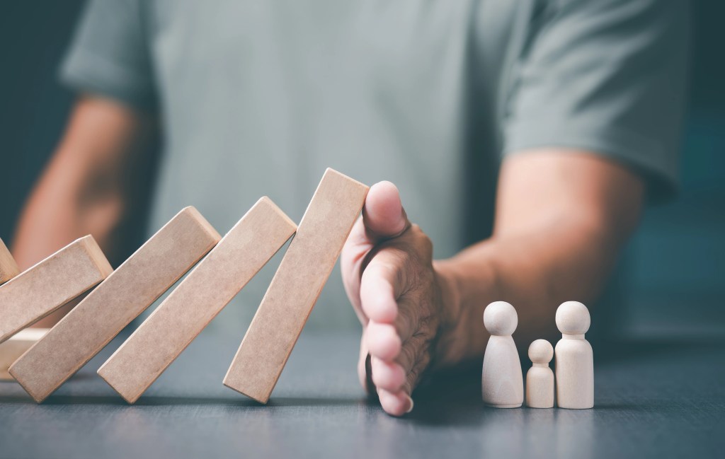 A hand stopping wooden blocks from falling, with three wooden figurines representing a family in the foreground.